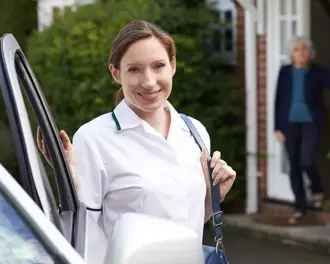  Smiling home carer in uniform arriving by personal vehicle, standing by the car outside a client’s house