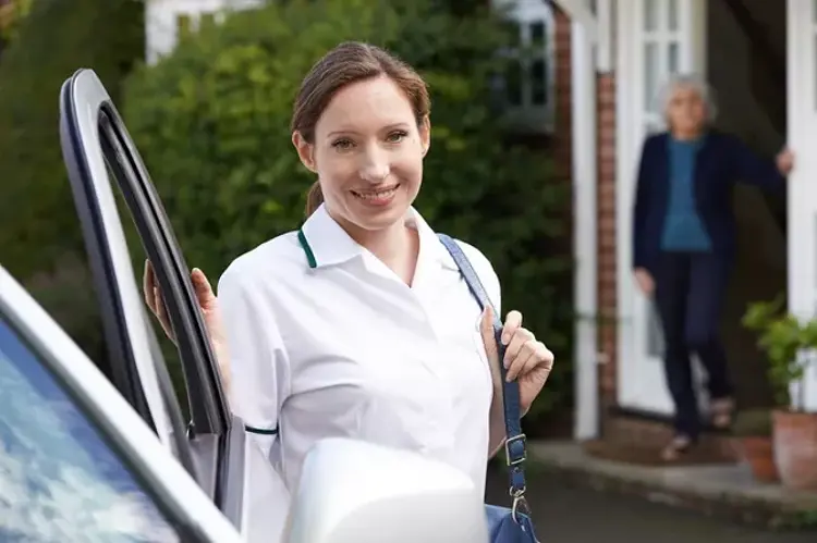 Smiling home carer in uniform arriving by personal vehicle, standing by the car outside a client’s house