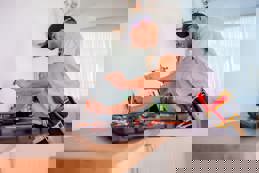 Handyman in a grey polo and black cap fixing plumbing equipment in a tidy kitchen with tools and metal pipes on the counter.