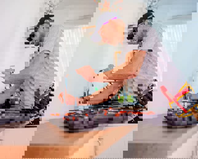 Handyman in a grey polo and black cap fixing plumbing equipment in a tidy kitchen with tools and metal pipes on the counter.