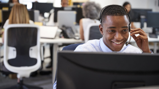A smiling man wearing a headset is working at a computer in a modern, open-plan office.