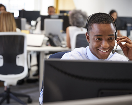 A smiling man wearing a headset is working at a computer in a modern, open-plan office.