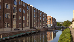 A canal with calm water reflecting brick apartment buildings on its left bank.