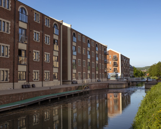A canal with calm water reflecting brick apartment buildings on its left bank.