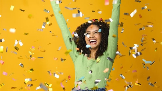A lady smiles against a yellow background as gold confetti falls around her