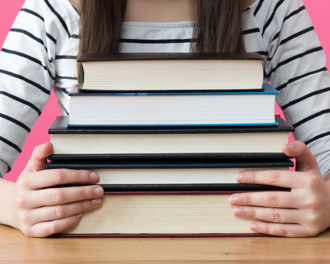 Student in striped shirt holding a stack of books on a desk against a pink background