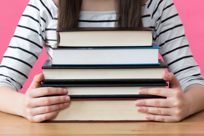 Student in striped shirt holding a stack of books on a desk against a pink background