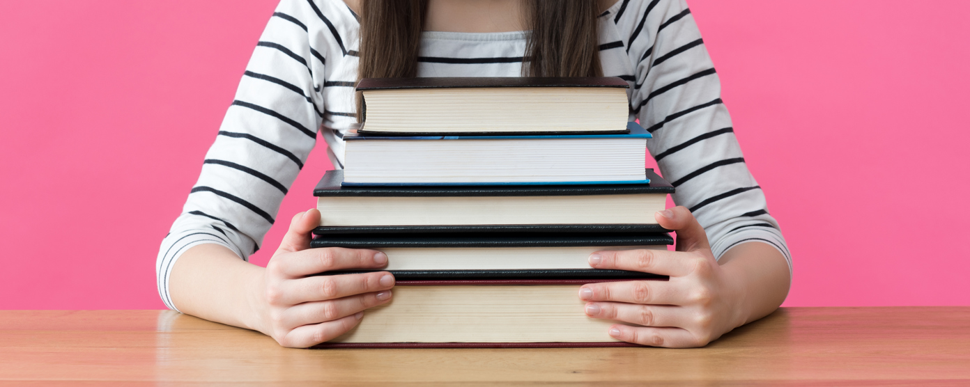 Student in striped shirt holding a stack of books on a desk against a pink background