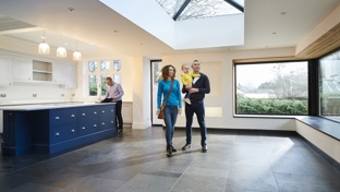 A young couple with a child tours a spacious, modern home with large windows and a skylight, while a real estate agent reviews documents.