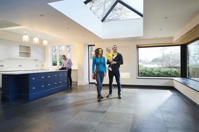 A young couple with a child tours a spacious, modern home with large windows and a skylight, while a real estate agent reviews documents.