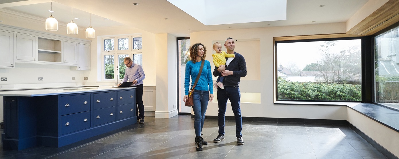 A young couple with a child tours a spacious, modern home with large windows and a skylight, while a real estate agent reviews documents.