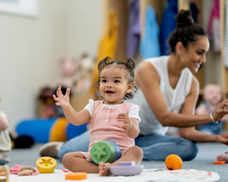 Toddlers sitting on a nursery floor with toys and an adult