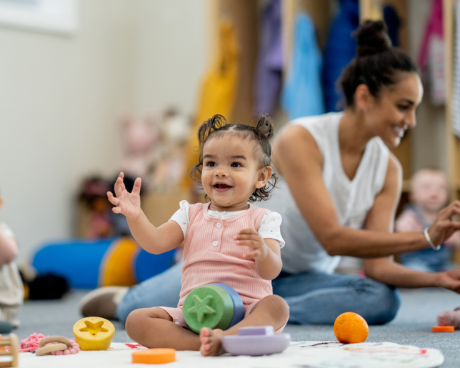 Toddlers sitting on a nursery floor with toys and an adult