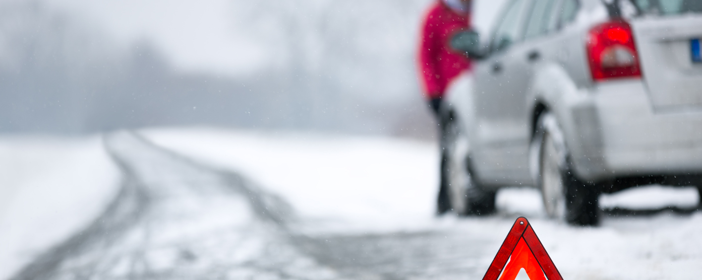 Red warning triangle on a snowy icy road with a broken-down car and driver in the background