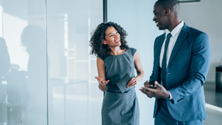 Two professionally dressed colleagues walking and chatting in a bright modern office corridor, smiling and engaged in conversation