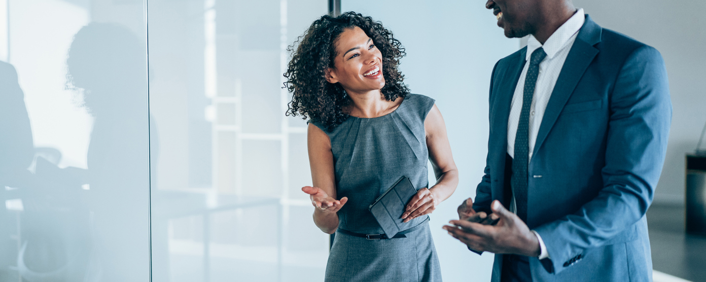 Two professionally dressed colleagues walking and chatting in a bright modern office corridor, smiling and engaged in conversation