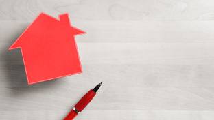 A red paper cutout in the shape of a house is positioned on the left side of a light woodgrain surface.
