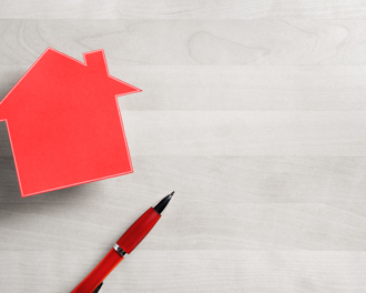 A red paper cutout in the shape of a house is positioned on the left side of a light woodgrain surface.
