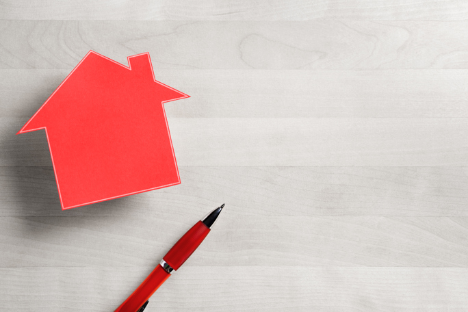 A red paper cutout in the shape of a house is positioned on the left side of a light woodgrain surface.