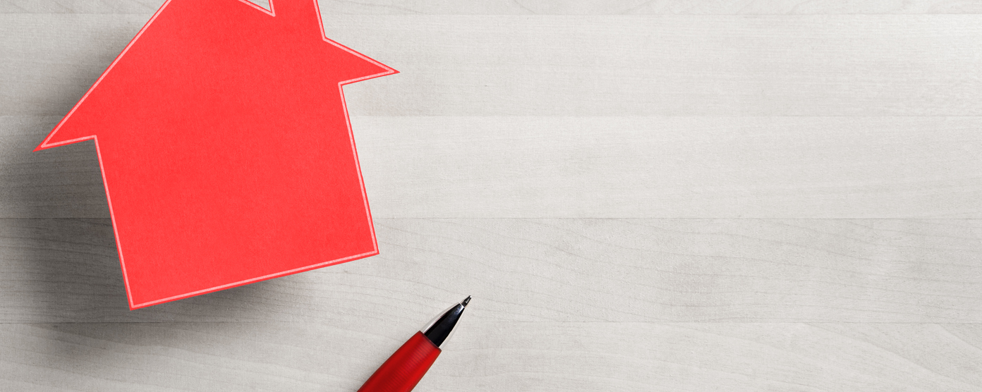 A red paper cutout in the shape of a house is positioned on the left side of a light woodgrain surface.