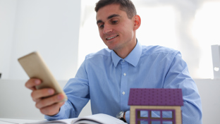 A man looks at a smartphone while seated at a desk, with a small model house positioned nearby.