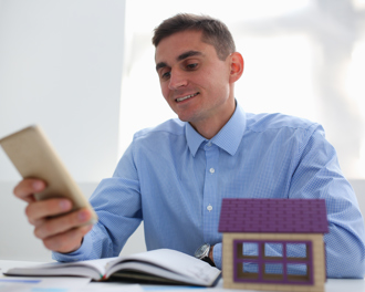 A man looks at a smartphone while seated at a desk, with a small model house positioned nearby.