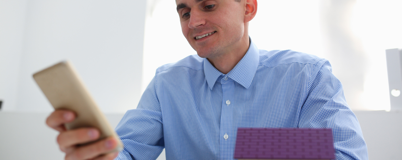 A man looks at a smartphone while seated at a desk, with a small model house positioned nearby.