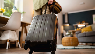 A person is seen from behind pulling a black hard-shell suitcase with wheels across the floor of a modern, cozy living room.