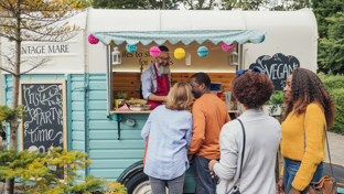  People queue at a colorful food truck, ordering from a bearded vendor at an outdoor market
