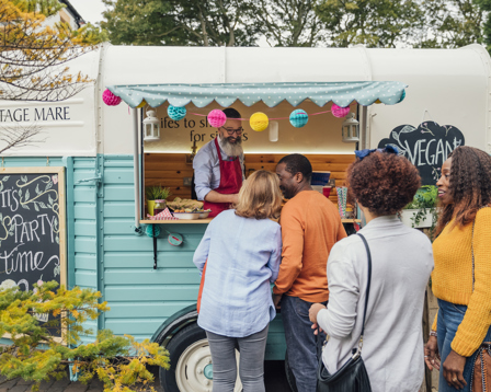  People queue at a colorful food truck, ordering from a bearded vendor at an outdoor market