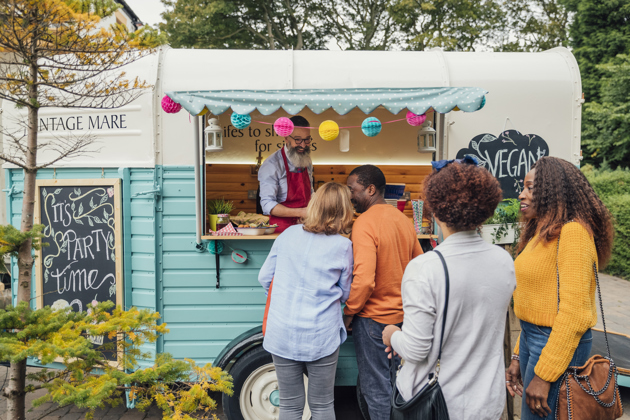  People queue at a colorful food truck, ordering from a bearded vendor at an outdoor market