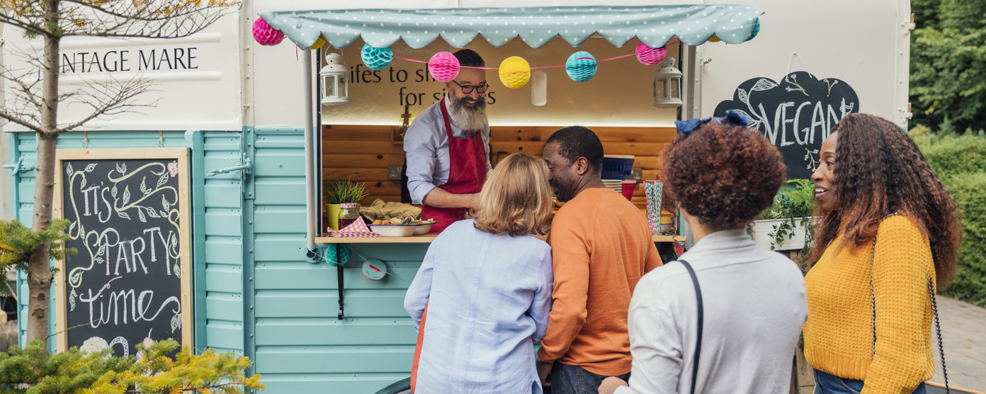  People queue at a colorful food truck, ordering from a bearded vendor at an outdoor market