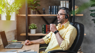 A man with a headset sits at a desk, focused on his laptop while holding a coffee cup, indicating a remote work environment.