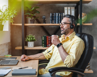 A man with a headset sits at a desk, focused on his laptop while holding a coffee cup, indicating a remote work environment.