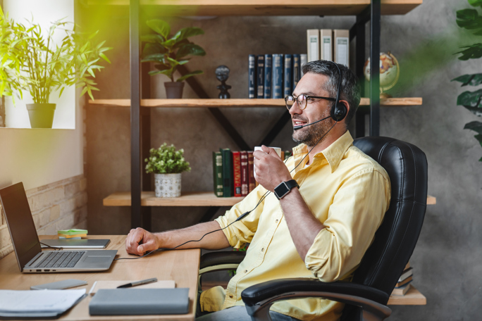 A man with a headset sits at a desk, focused on his laptop while holding a coffee cup, indicating a remote work environment.