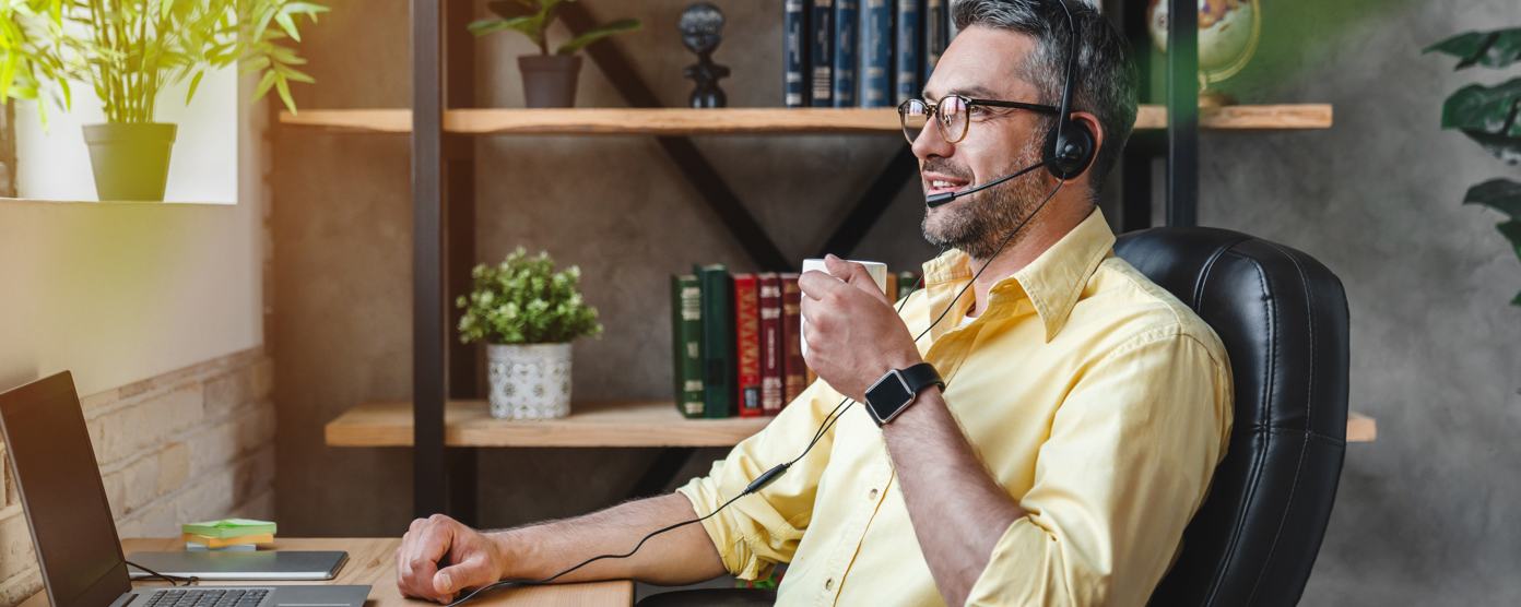 A man with a headset sits at a desk, focused on his laptop while holding a coffee cup, indicating a remote work environment.