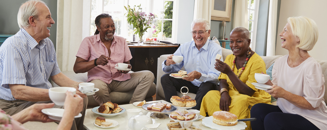 Group of senior friends laughing and enjoying afternoon tea with cakes in a bright living room