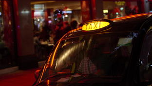 Close-up of a London black cab with the taxi sign lit, reflecting red neon lights outside a busy venue at night