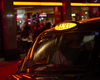 Close-up of a London black cab with the taxi sign lit, reflecting red neon lights outside a busy venue at night