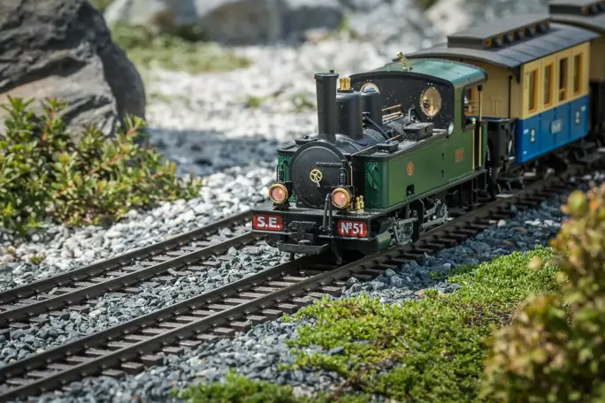 A green and black model train on a gravel track, with green foliage and rocks in the background