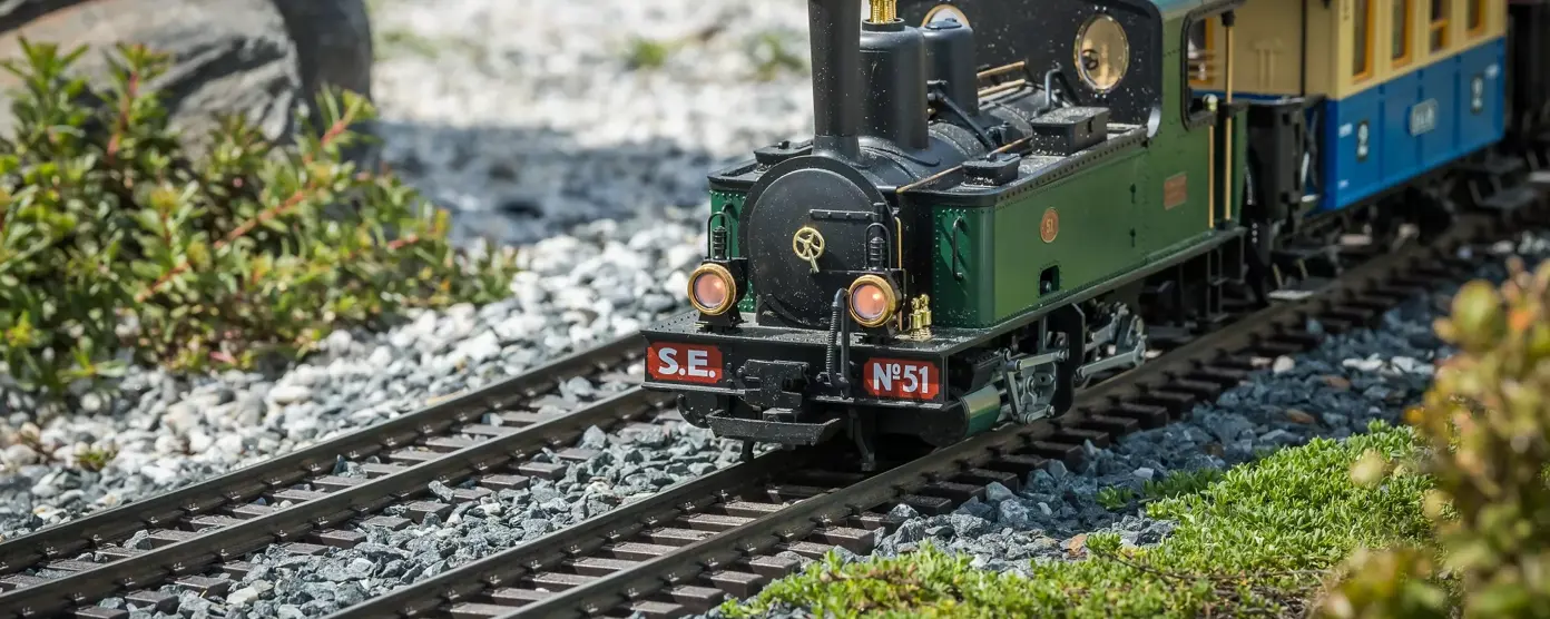 A green and black model train on a gravel track, with green foliage and rocks in the background