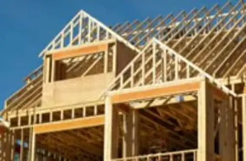 A wooden house frame under construction, with slanted roof beams and a clear blue sky in the background