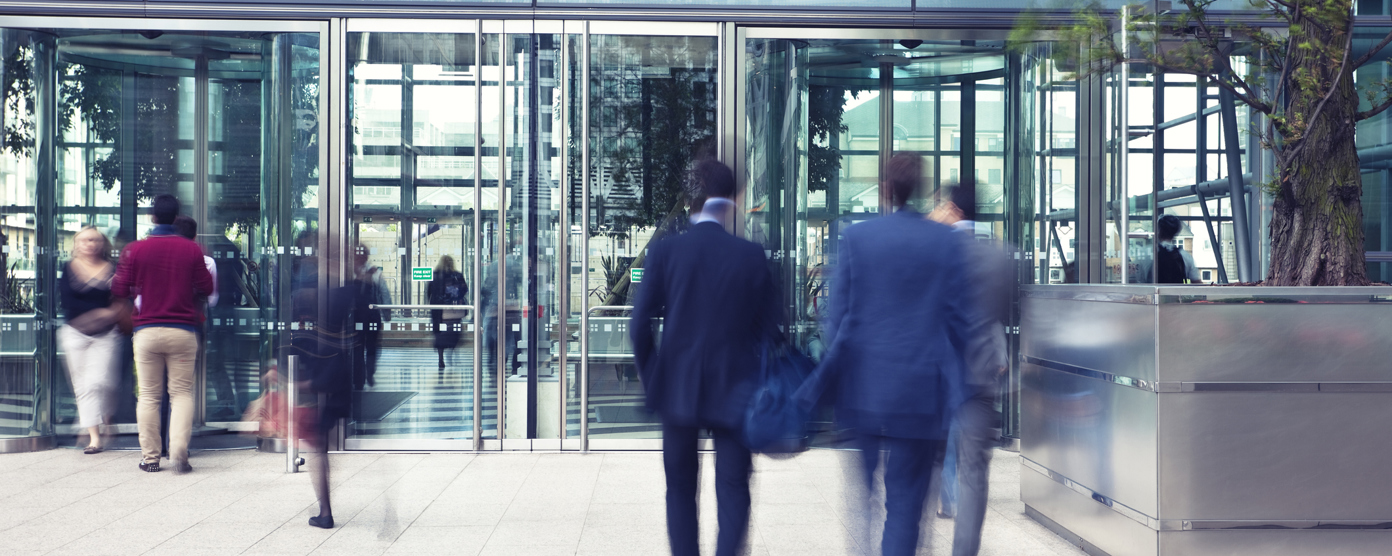 Business professionals actively entering and exiting a modern office building with a sleek glass facade, showcasing corporate life.