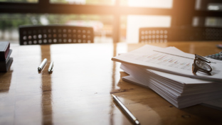A close-up image of a wooden conference table with several pens and a large stack of documents.