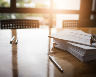 A close-up image of a wooden conference table with several pens and a large stack of documents.