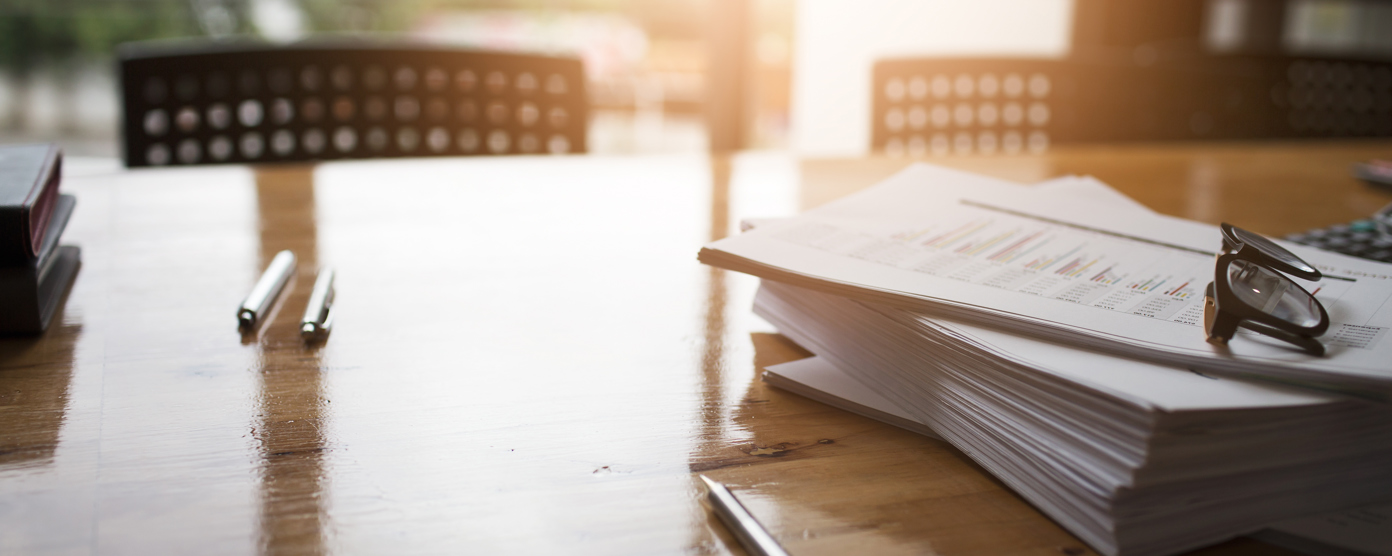 A close-up image of a wooden conference table with several pens and a large stack of documents.