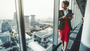 Professional businesswoman with tablet standing by office window overlooking cityscape.
