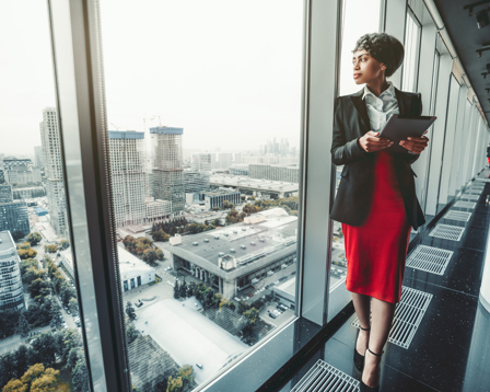 Professional businesswoman with tablet standing by office window overlooking cityscape.