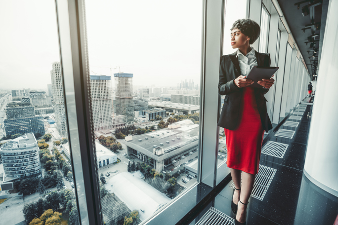 Professional businesswoman with tablet standing by office window overlooking cityscape.