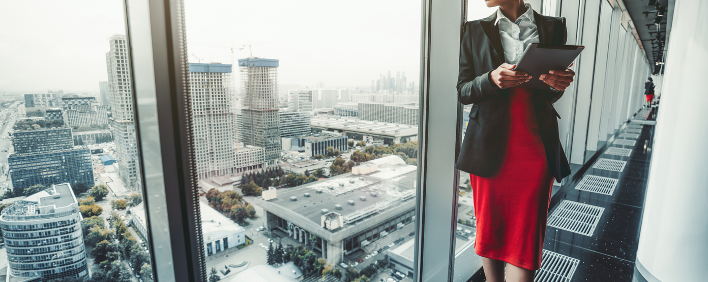 Professional businesswoman with tablet standing by office window overlooking cityscape.